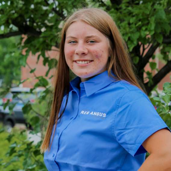 Young woman with long brown hair wearing a blue "Red Angus" shirt stands outside in front of green foliage, smiling at the camera.