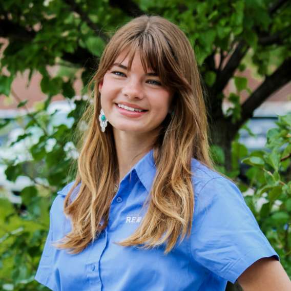 A young woman with long brown hair wearing a blue collared shirt stands outdoors in front of green leafy trees, smiling at the camera.