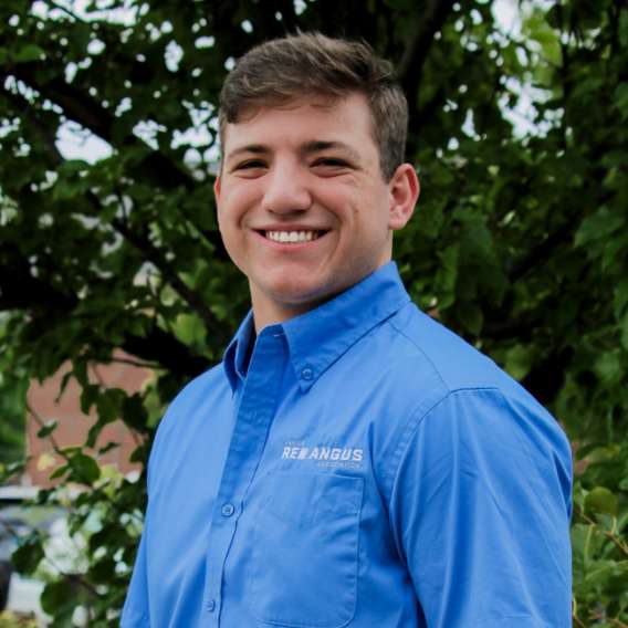 A person wearing a blue button-up shirt with "Red Angus" text on the pocket stands outside in front of green foliage, smiling at the camera.