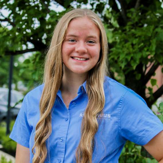 A young woman with long blonde hair wearing a light blue button-up shirt stands outdoors, smiling in front of green foliage.