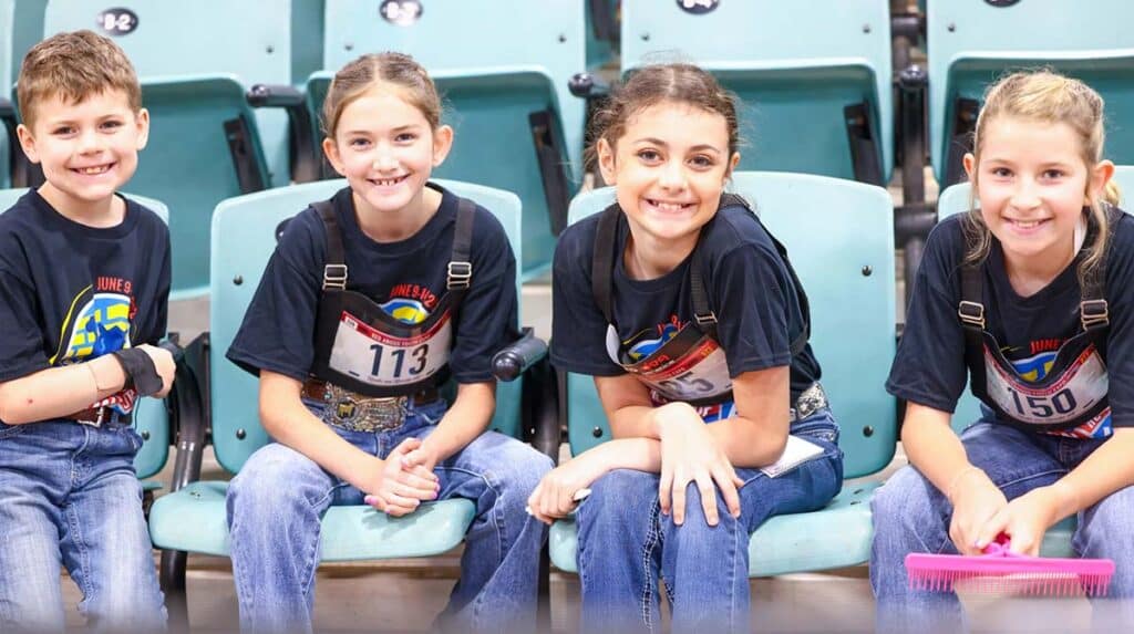 Four children wearing matching shirts and jeans sit on teal seats, smiling at the camera, each with a number tag pinned to their shirts.