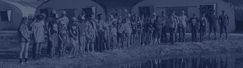 A large group of people stands in a line outdoors near a pond, with several industrial buildings visible in the background.