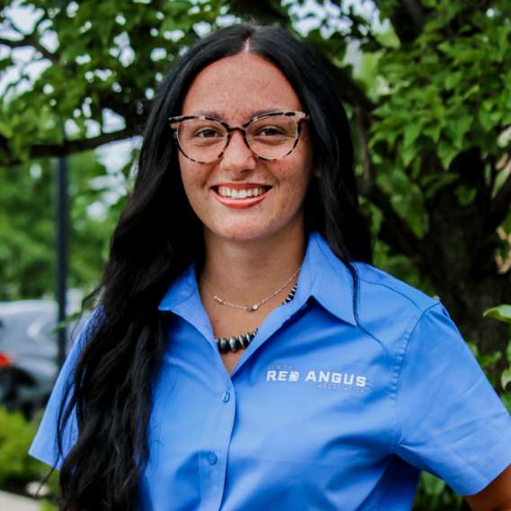 A person with long dark hair and glasses smiles outdoors, wearing a blue collared shirt with "RED ANGUS" embroidered on it. Green foliage is visible in the background.
