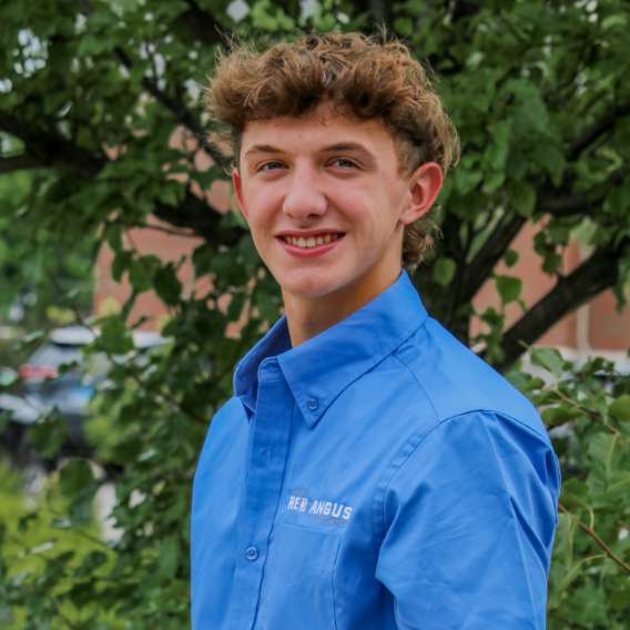 A young person with curly hair wearing a blue "Red Angus" shirt stands outdoors in front of a leafy tree, smiling at the camera.