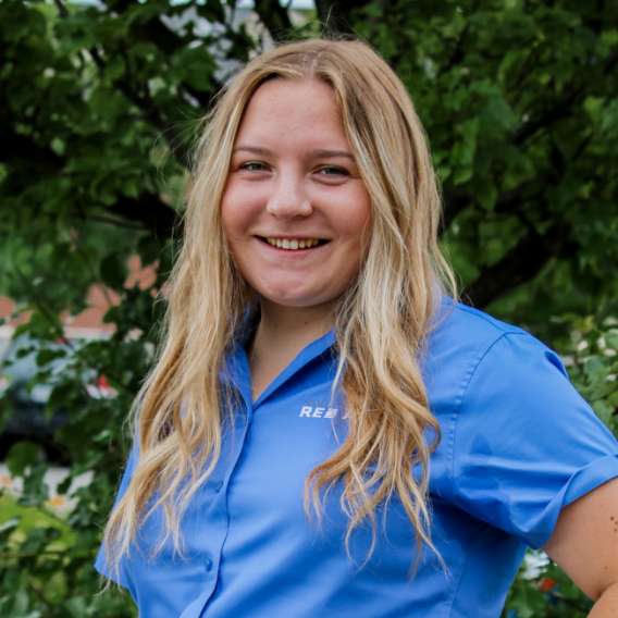 A person with long blonde hair wearing a blue collared shirt stands outdoors in front of green foliage, smiling at the camera.