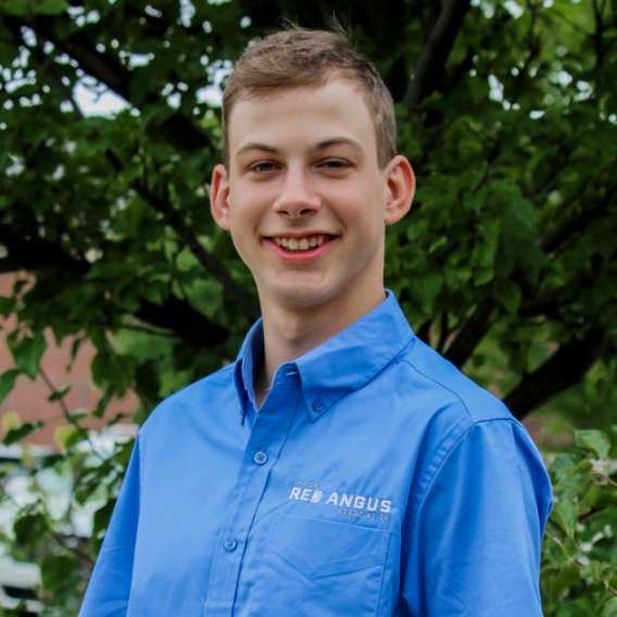 A young man in a blue "Red Angus" shirt stands outdoors in front of green leafy trees, smiling at the camera.